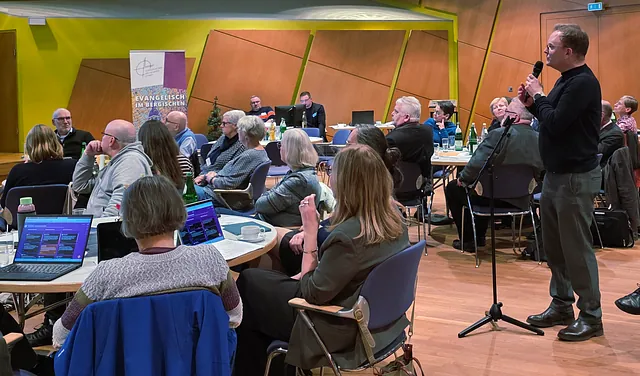 Blick in den Tagungsraum des Kresynode des Ev. Kirchenkreises mit vielen Menschen an den Tischen und Laptops darauf. Ein Mann steht am Mikrohalter und hält das Mikrophon in den Händen.