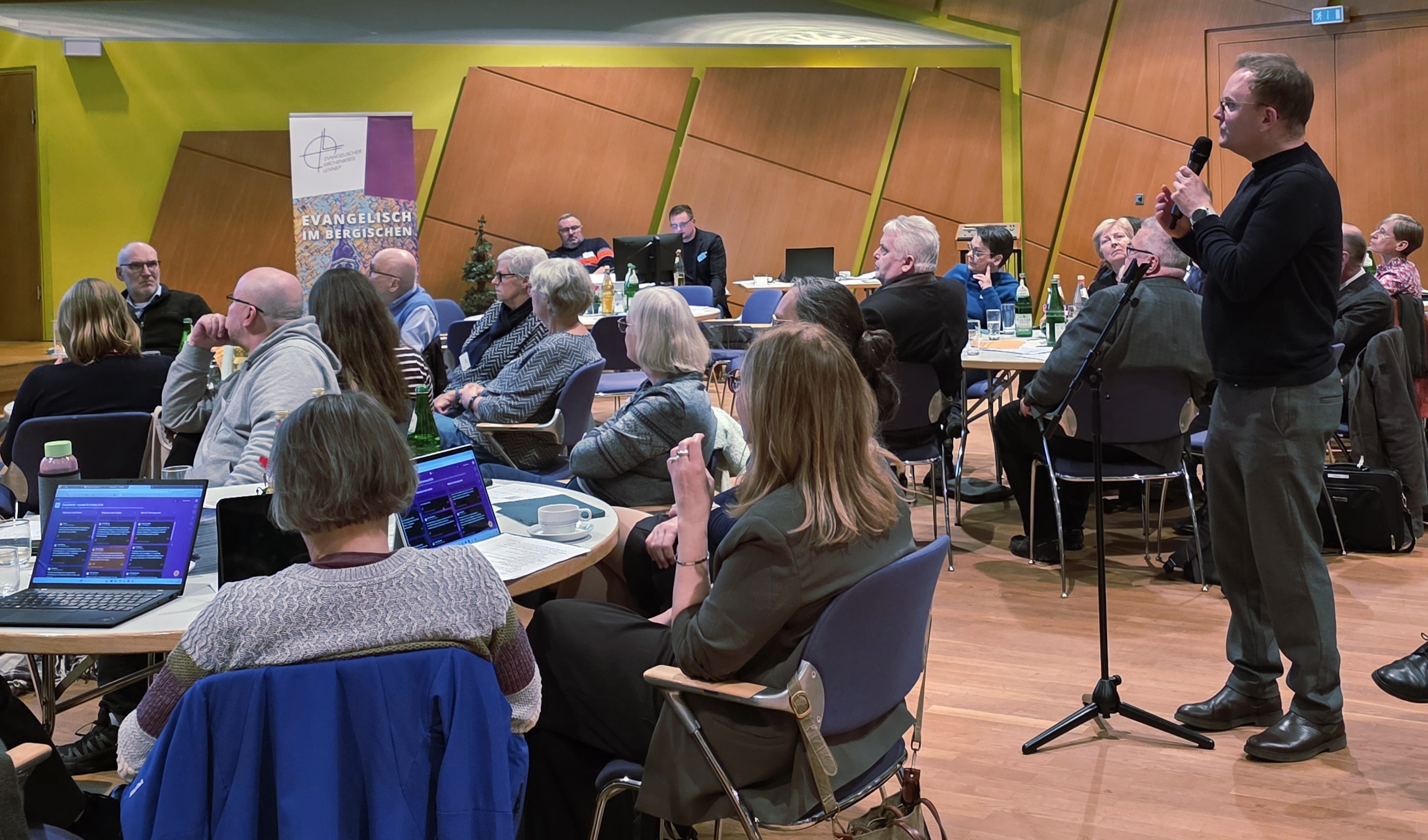 Blick in den Tagungsraum des Kresynode des Ev. Kirchenkreises mit vielen Menschen an den Tischen und Laptops darauf. Ein Mann steht am Mikrohalter und hält das Mikrophon in den Händen.