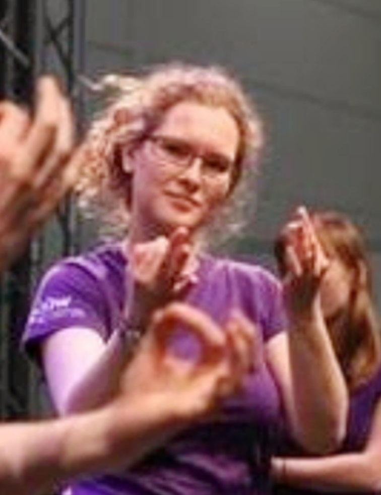 Frau mit Locken und Brille im lila Shirt, umgeben von unscharfen „Okay“-Gesten.