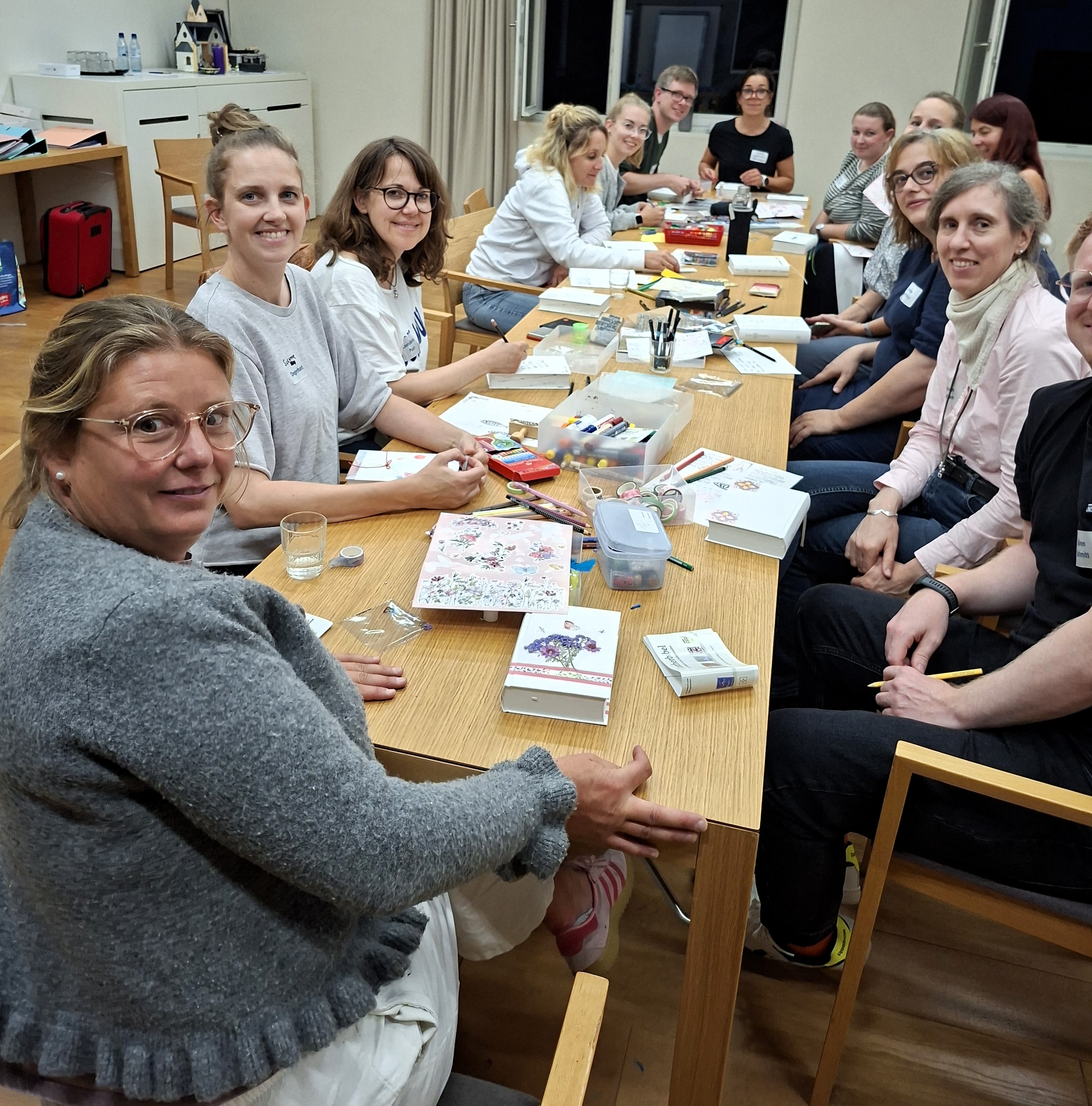 Menschen sitzen an einem Tisch und schauen in die Kamera. Auf dem Tisch dicke weiße Bücher und Stifte sowie Bastelmaterialien. Stifte 
