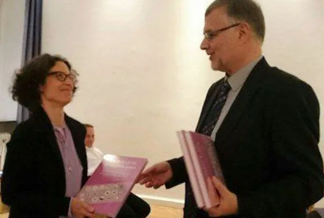 Frau mit Locken und Brille erhält lila Buch von lächelndem Mann im Anzug.