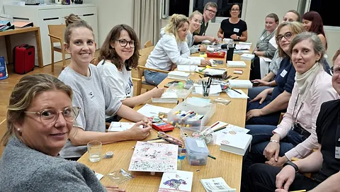 Menschen sitzen an einem Tisch und schauen in die Kamera. Auf dem Tisch dicke weiße Bücher und Stifte sowie Bastelmaterialien. Stifte 