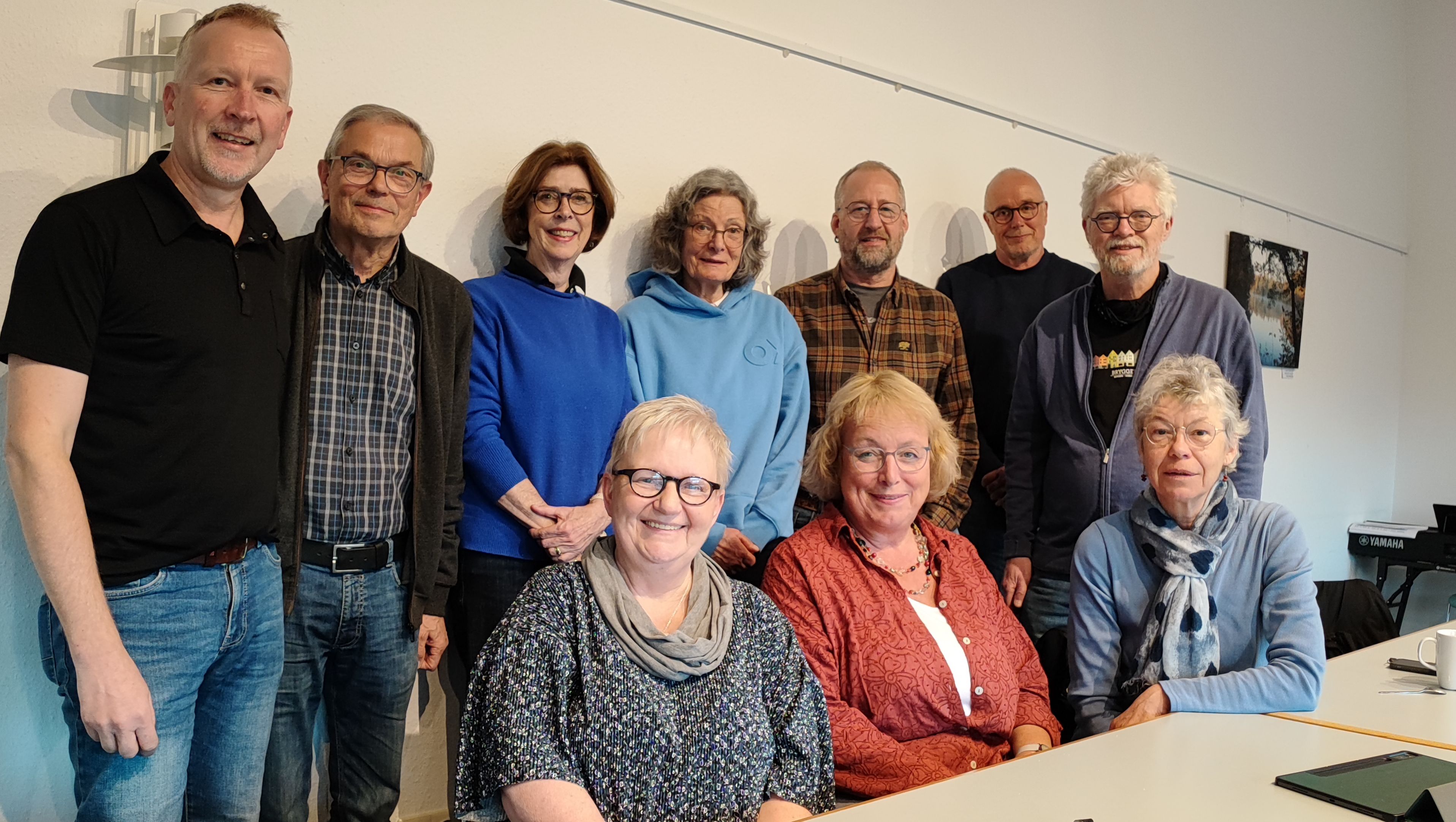 Zehn Menschen lächeln in die Kamera, drei von ihnen sitzen. Sie sind älter. Vor ihnen stehen Kaffeetassen auf dem Tisch.