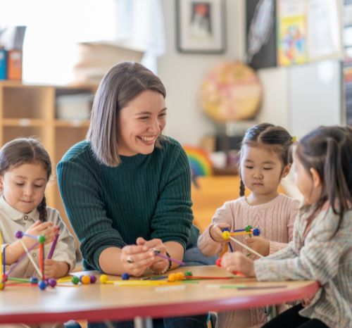Eine junge Frau sitzt lachend mit Kindern am Tisch während sie Stifte halten