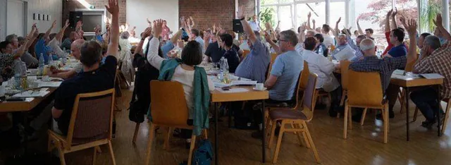 Panoramablick auf Versammlung, viele heben die Hand.