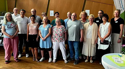 Viele Menschen stehen in sommerlicher Kleidung vor einer Holzwand und lächeln. 
